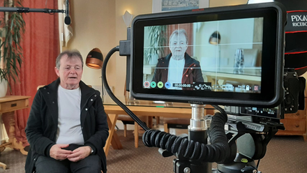 Photo of an interviewee seen both on a monitor on the right of the screen and in person on the left.