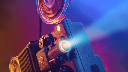 Atmospheric image of an old film projector with its white light beam cutting through a purple and blue hazed dark room.