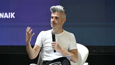 Evan Shapiro speaking on stage at Stream TV. He is wearing a white t-shirt and signalling with his hands in front of a purple background.