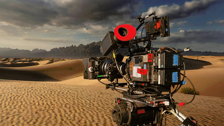 A large broadcast camera sits sits atop a tripod, looking out over a vast empty desert landscape.