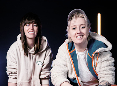 Two women pose in light hoodies in a dark room with white bar neon lights in the background.