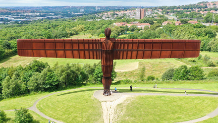 The Angel of the North monument stands large in the foreground atop a grassy mound, with buildings and trees visible in the background behind it.