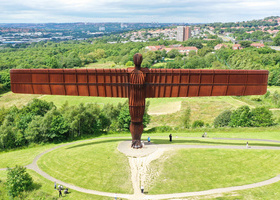 The Angel of the North monument stands large in the foreground atop a grassy mound, with buildings and trees visible in the background behind it.