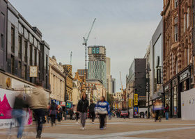 Briggate, Leeds; a busy pedestrian street in Leeds city centre, lined with shops and buildings with cranes in the background. 