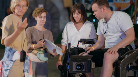 Four crew members stand behind a large camera on a tripod, discussing the scene they are about to shoot.