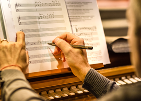 Close up of hands writing on a music sheet which is propped up on a piano or organ.