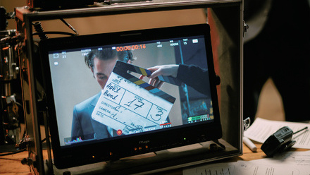 Photo of a screen on a desk with an image of an actor with a clapper board obstructing his face. There is a walkie talkie and script pages beside it.