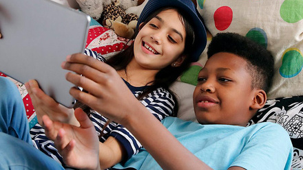 Two children lying down on a colourful bed holding a tablet above their heads.