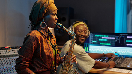 Musician playing a saxophone in a recording studio while a sound engineer adjusts audio tracks at a mixing console.