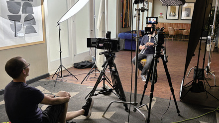A producer/director sits behind a two camera interview set-up, with an interviewee seated in front of the cameras but obscured from view.