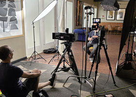 A producer/director sits behind a two camera interview set-up, with an interviewee seated in front of the cameras but obscured from view.