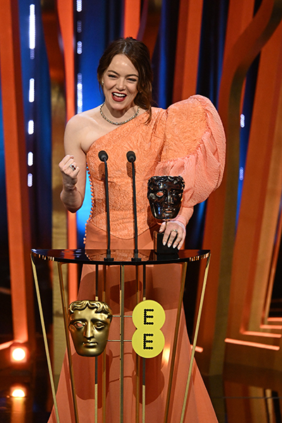 Photo of actress Emma Stone, a white woman with dark red hair, on stage with her BAFTA award, stood behind a podium and fist pumping the air in celebration. She is smiling.