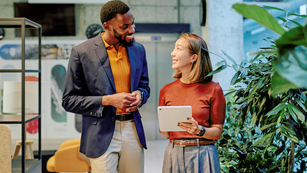 Two workers walking through a modern green office, discussing project details while using a tablet in a bright, airy workspace. 