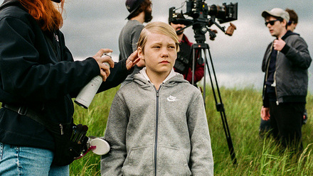 A child actor having their hair sprayed by a hair stylist on set in a rural filming location with camera crew and equipment in the background.