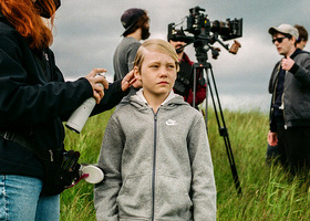 A child actor having their hair sprayed by a hair stylist on set in a rural filming location with camera crew and equipment in the background.