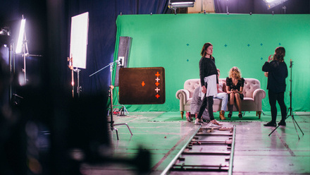 A small group of actors and production crew wait between takes in front of a large green screen in a studio.