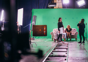 A small group of actors and production crew wait between takes in front of a large green screen in a studio.