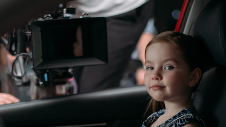 A little girl with blond hair sitting in a car seat looks to the camera, in the background is a large lens of a video camera resting on the car window