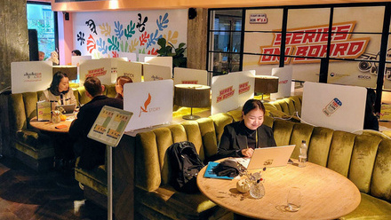 A Korean person sits at a round table with their laptop open. Behind them is a large sign which says 'Series on Board'.