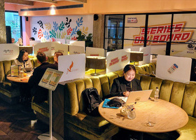 A Korean person sits at a round table with their laptop open. Behind them is a large sign which says 'Series on Board'.