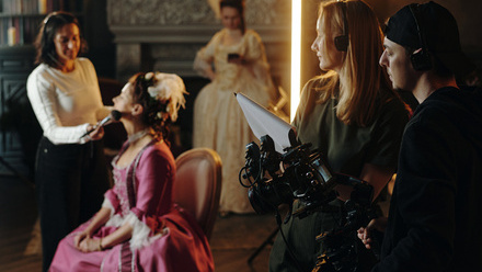 Behind-the-scenes on a period drama where a makeup artist is touching up an actor and two crew members stand nearby with a camera.