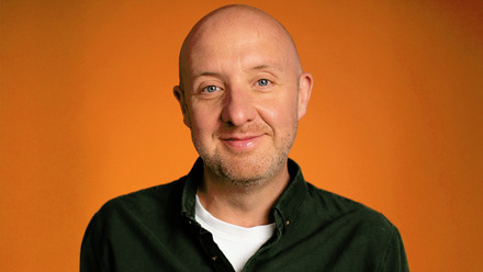 Headshot: Daniel Nettleton, a bald man wearing a white t-shirt underneath a collared shirt in front of an orange background.