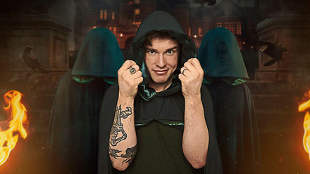 Ed Gamble posing in a Traitors cloak with raised fists in a dark, fantasy setting with flames and shadowy figures behind him.