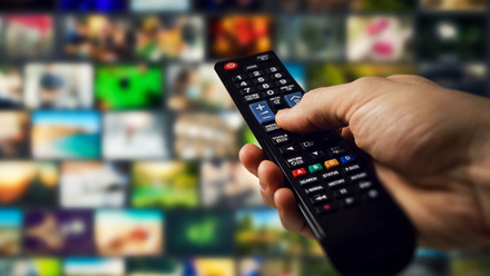 Close-up of a hand holding a TV remote control, pointing at a screen filled with multiple streaming content thumbnails.