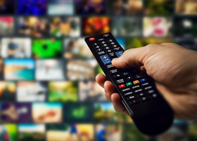 Close-up of a hand holding a TV remote control, pointing at a screen filled with multiple streaming content thumbnails.