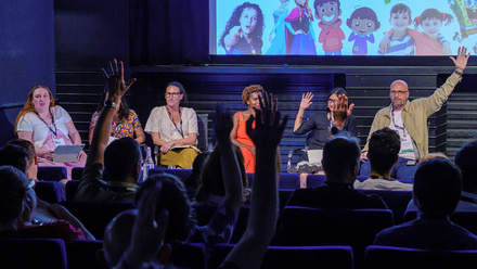 A panel of speakers on stage at the Children's Media Conference sat in front with an audience of people raising their hands.
