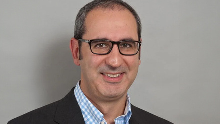 Headshot: Sammy Nourmand, a man with very short dark hair wearing black glasses, a black suit jacket and light blue and white checked shirt.