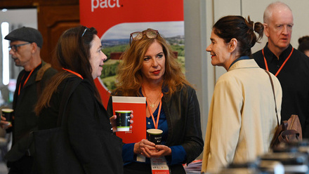 Three women stand talking in front of a red Pact pop-up banner, with other delegates visible in the background.