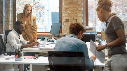 A glass walled office with a diverse team of employees sitting at and standing near desks with computers and tablets.