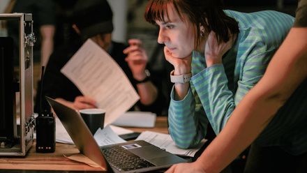 Photo of a woman with short brown hair on a set, crouched down over a laptop whilst a man behind her is reviewing pages of a script with a pen.