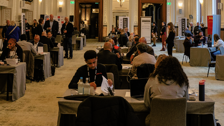 Photo of a large traditional ballroom filled with tables in a row where small meetings are taking place amongst lots of groups of people.