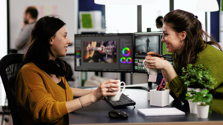Photo image of two women smiling and chatting in an office while holding mugs, with video editing software open on their computer screens.