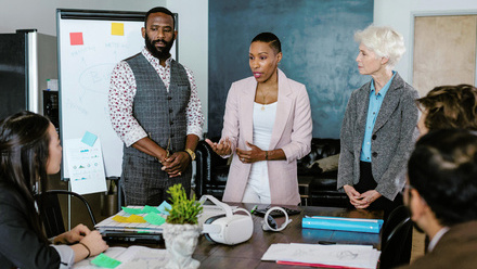 Team meeting in an office setting. Three people are stood up at a table wearing smart outfits, three others are sat down listening.
