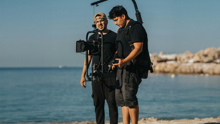 Camera operator with a stabiliser stood next to a film crew member in a coastal location. 