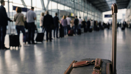 A suitcase sits in a large airport terminal with a glass wall; a long queue of people with luggage can be seen in the background.