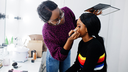 Photo image of two black women in a makeup room, one is sat in front of the mirror whilst the other applies makeup to her eyelid.