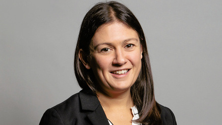 Headshot: Lisa Nandy, a woman with dark shoulder-length hair wearing a white shirt and black suit jacket, stood in front of a grey backdrop.