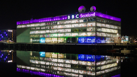 A large glass building at night lit up in white and purple light, with 'BBC Scotland' written in large letters across the top floor.