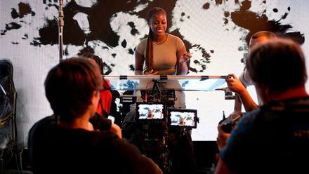 A person speaks on set while a film crew records them against a black-and-white abstract backdrop.