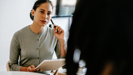Photo of a woman in an office environment, she is listening intently to someone say opposite her and is holding a pen and a pad of paper.