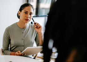 Photo of a woman in an office environment, she is listening intently to someone say opposite her and is holding a pen and a pad of paper.