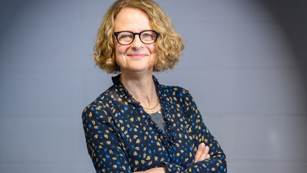 Headshot: Ruth Pitt, a woman with a curly fair bob wearing glasses and a floral print blue and yellow shirt.