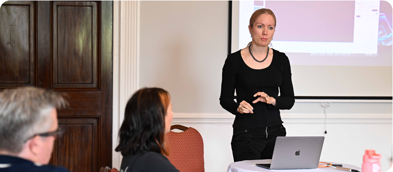A guest speaker delivers a talk whilst workshop participants listen, the corner of a projector screen is visible behind the speaker.