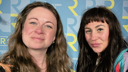 Two women with long hair smile to the camera, stood in front of a large step and repeat board in blue with an 'R' motif at Realscreen Summit.