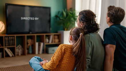 A family of three sit on a sofa facing a large TV with the words 'DIRECTED BY' on the screen.