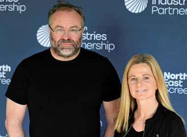 Duncan Gray and Antonia Hurford-Jones, both dressed in black tops, smile for the camera stood in front of a blue North East Screen branding board.